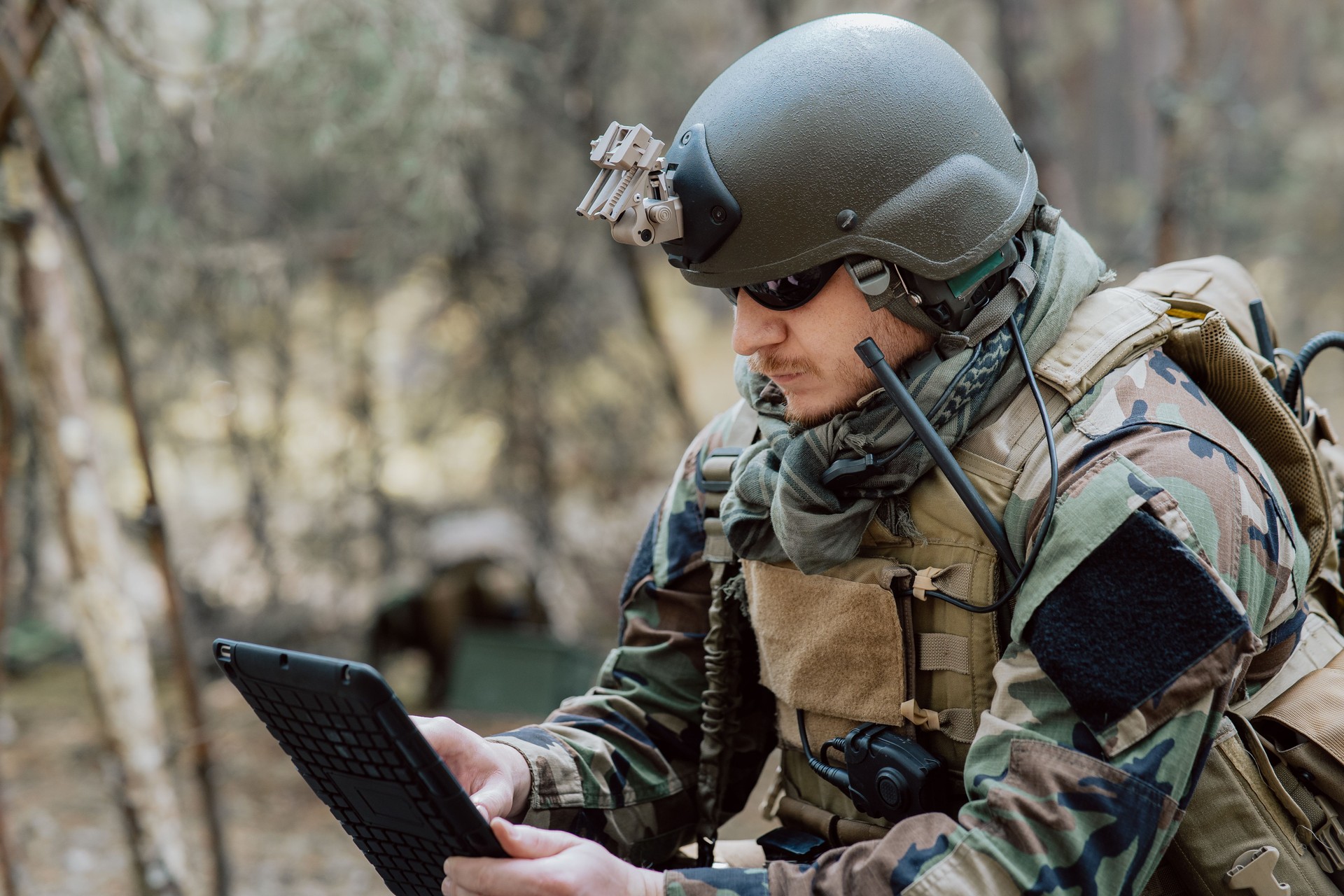 Bearded soldier in uniform sit on military transport crates, analyze data on a tablet and work out tactics at a temporary forest base. In the background, you can see a soldier protecting the base